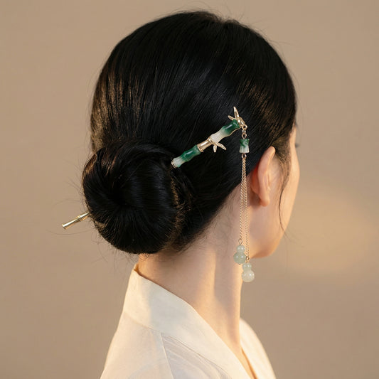 Rear view of a woman with jet-black hair in an elegant low bun at the nape, secured with a gold and jade-green enamel bamboo hair stick, its gold chain and jade gourd pendant draping gracefully against her hair