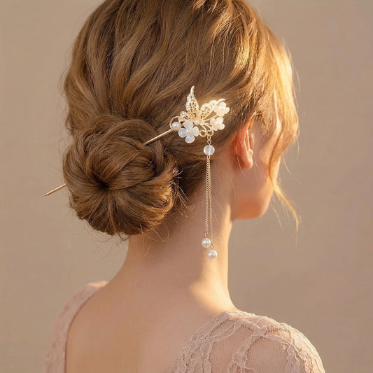 Rear view of a woman with honey-brown wavy hair in a low bun secured with a gold butterfly and opalescent flower hair stick, its pearl chain draping beside her ear, wearing a delicate lace top